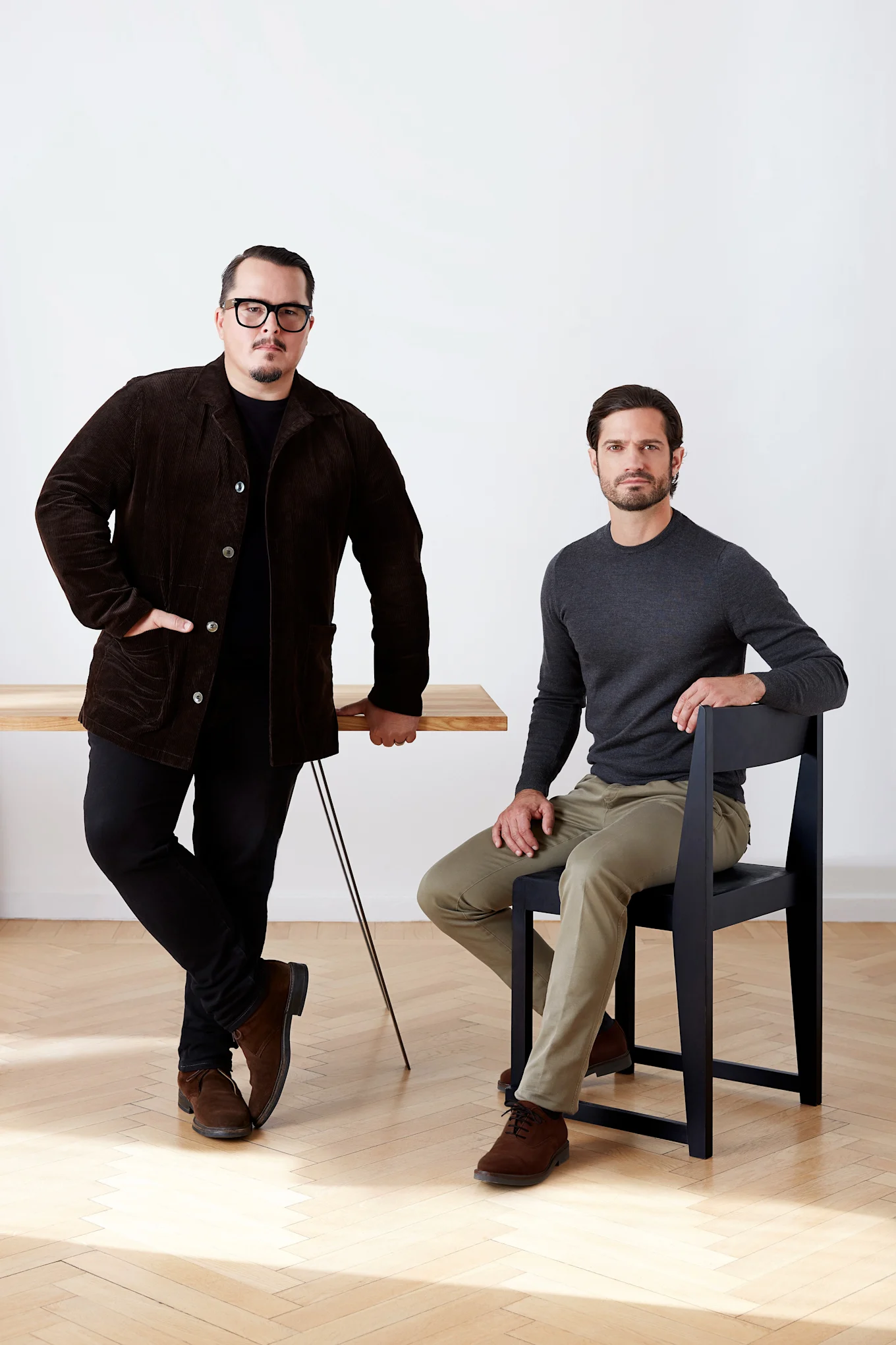 To mænd, den ene står ved et træbord i en brun fløjlsjakke, den anden sidder på en sort stol i en grå skjorte.