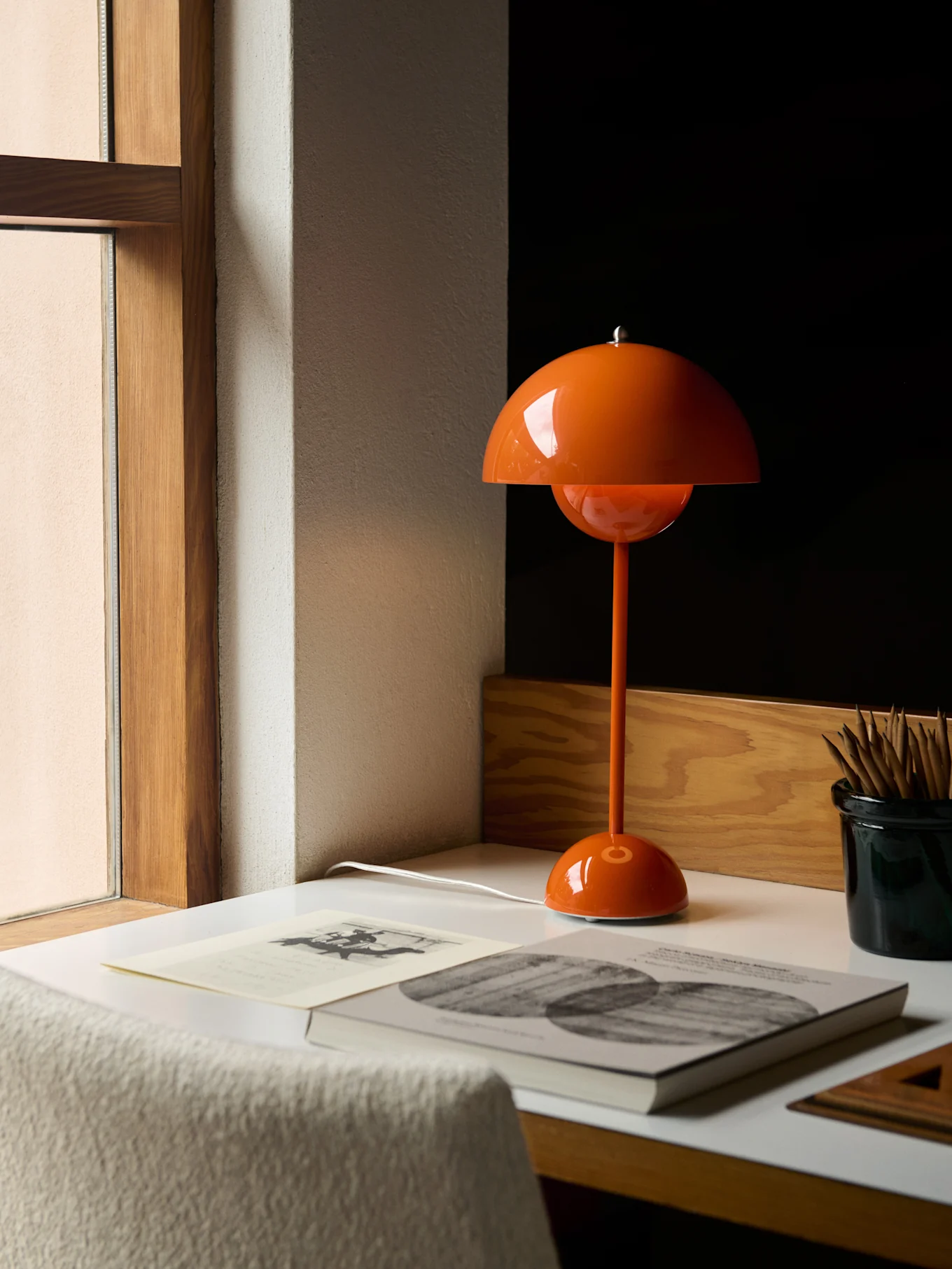 En orange, champignonformet bordlampe på et hvidt skrivebord ved siden af et vindue, med bøger og blyanter.