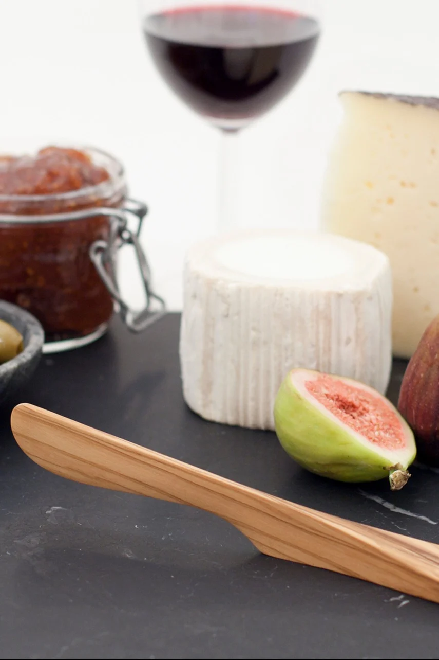Et ostebord med en trækniv, to oste, en halveret figen, marmelade, oliven og et glas rødvin.