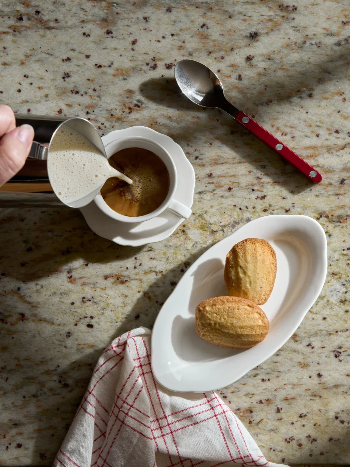 Hånd hælder skummet mælk i en kaffekop ved siden af madeleinekager på en granitbordplade.
