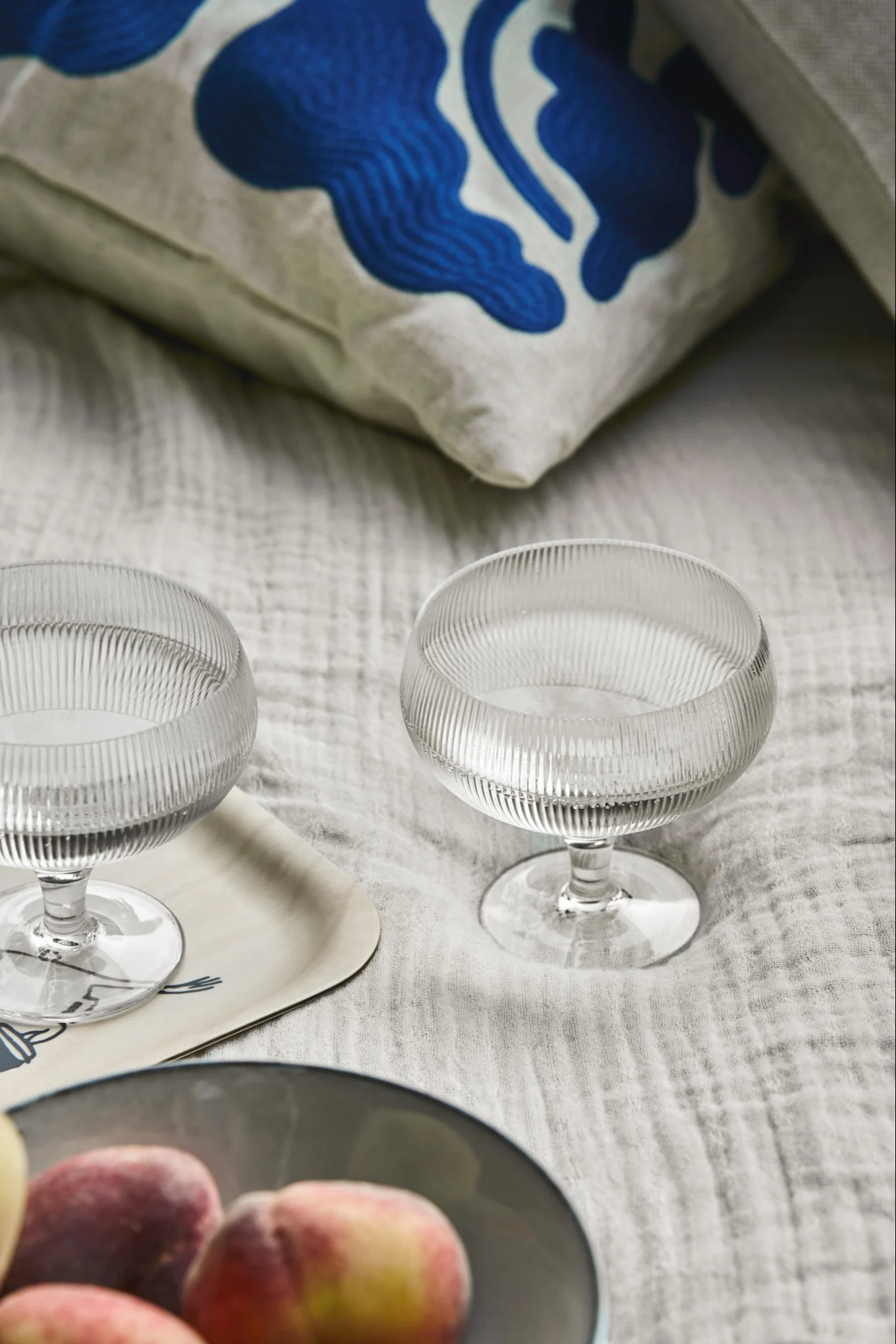 To riflede coupeglas på en lys bakke, med ferskner i en skål og en blåmønstret pude i baggrunden.