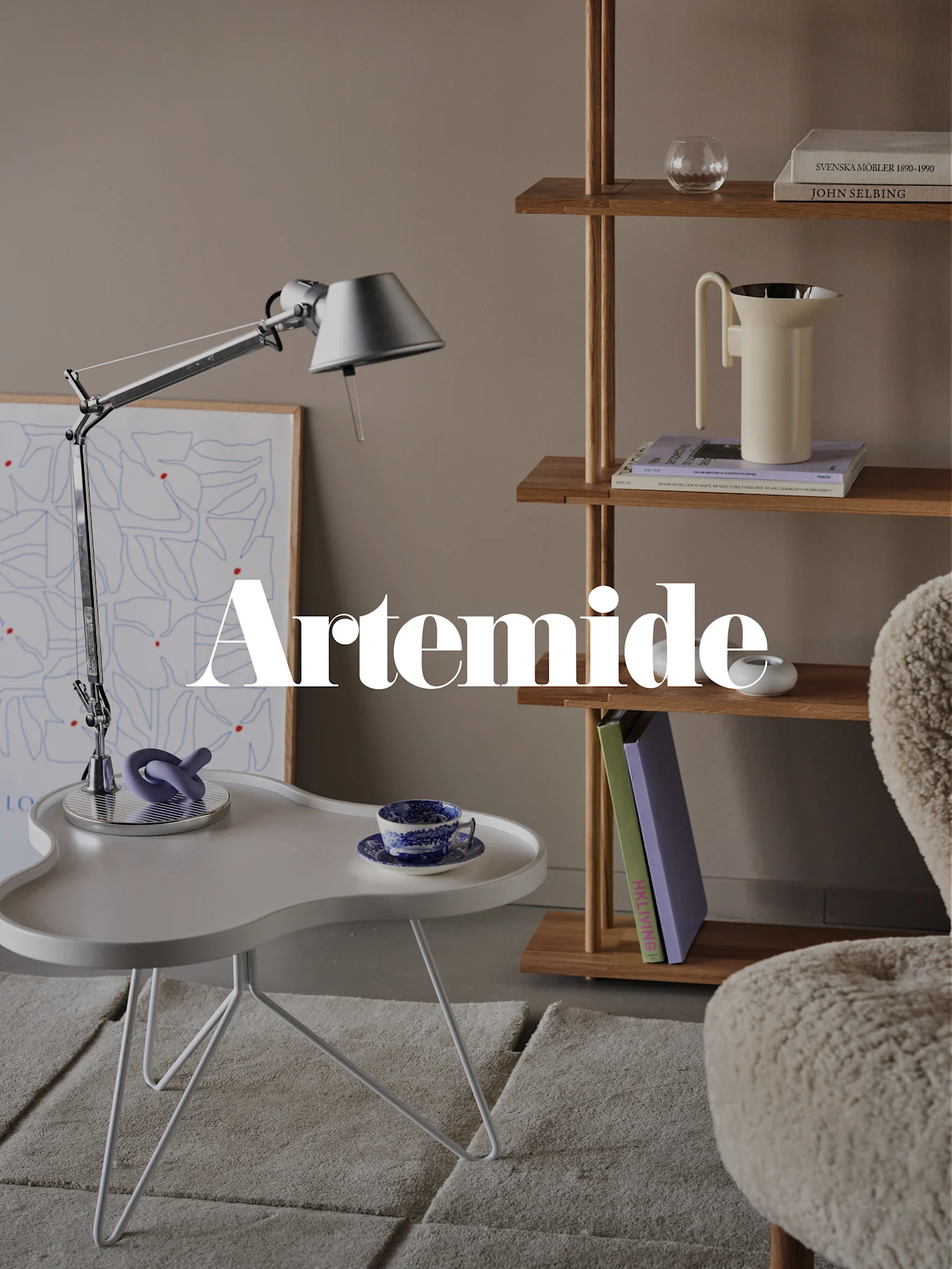 En moderne stue med en sølvfarvet bordlampe, hvidt sidebord, træbogreol og en cremefarvet lænestol. Tekstoverlejring: 'Artemide'.