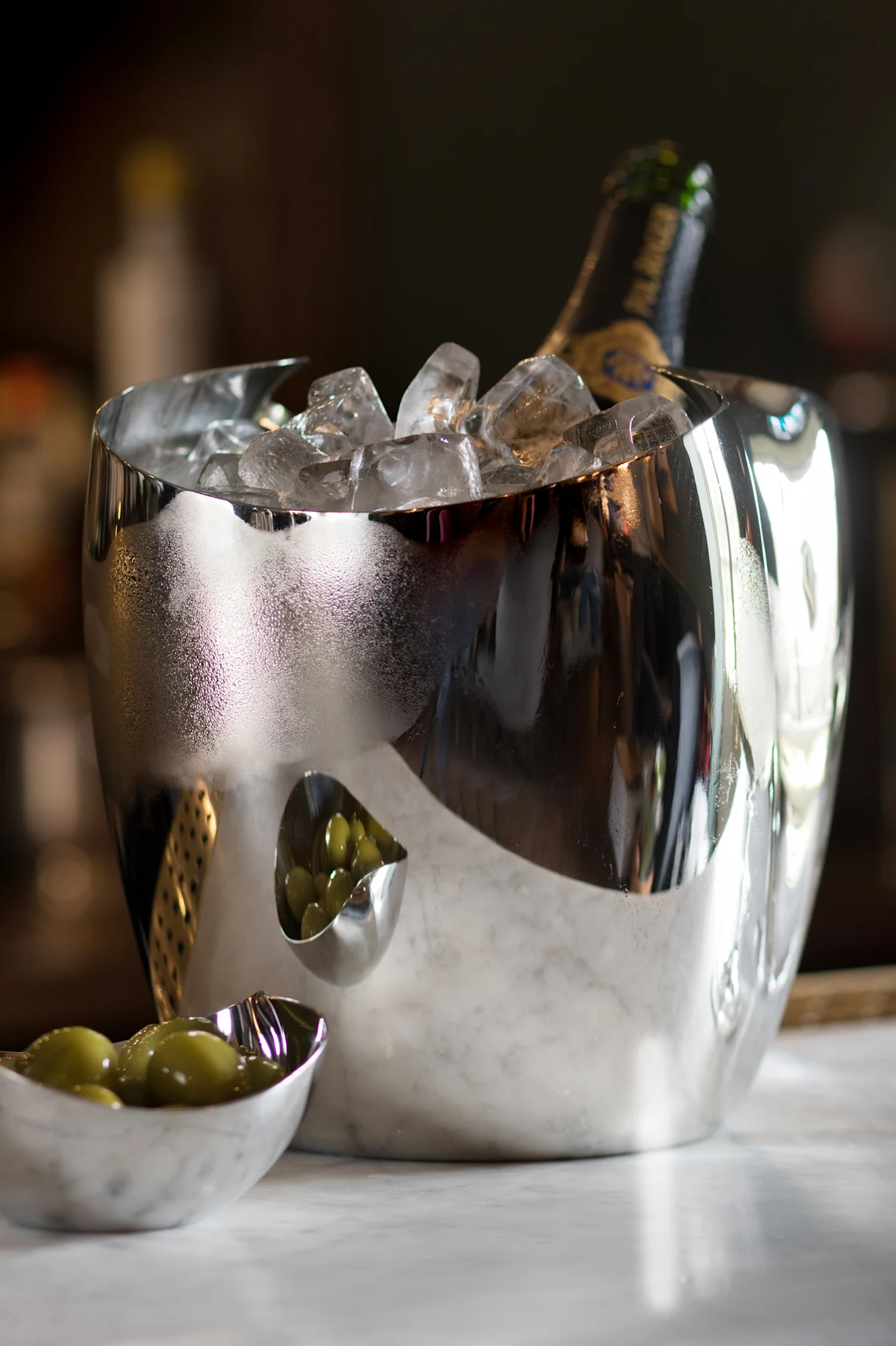 En reflekterende sølv isspand med en champagneflaske og is, ved siden af to små skåle med grønne oliven på marmor.