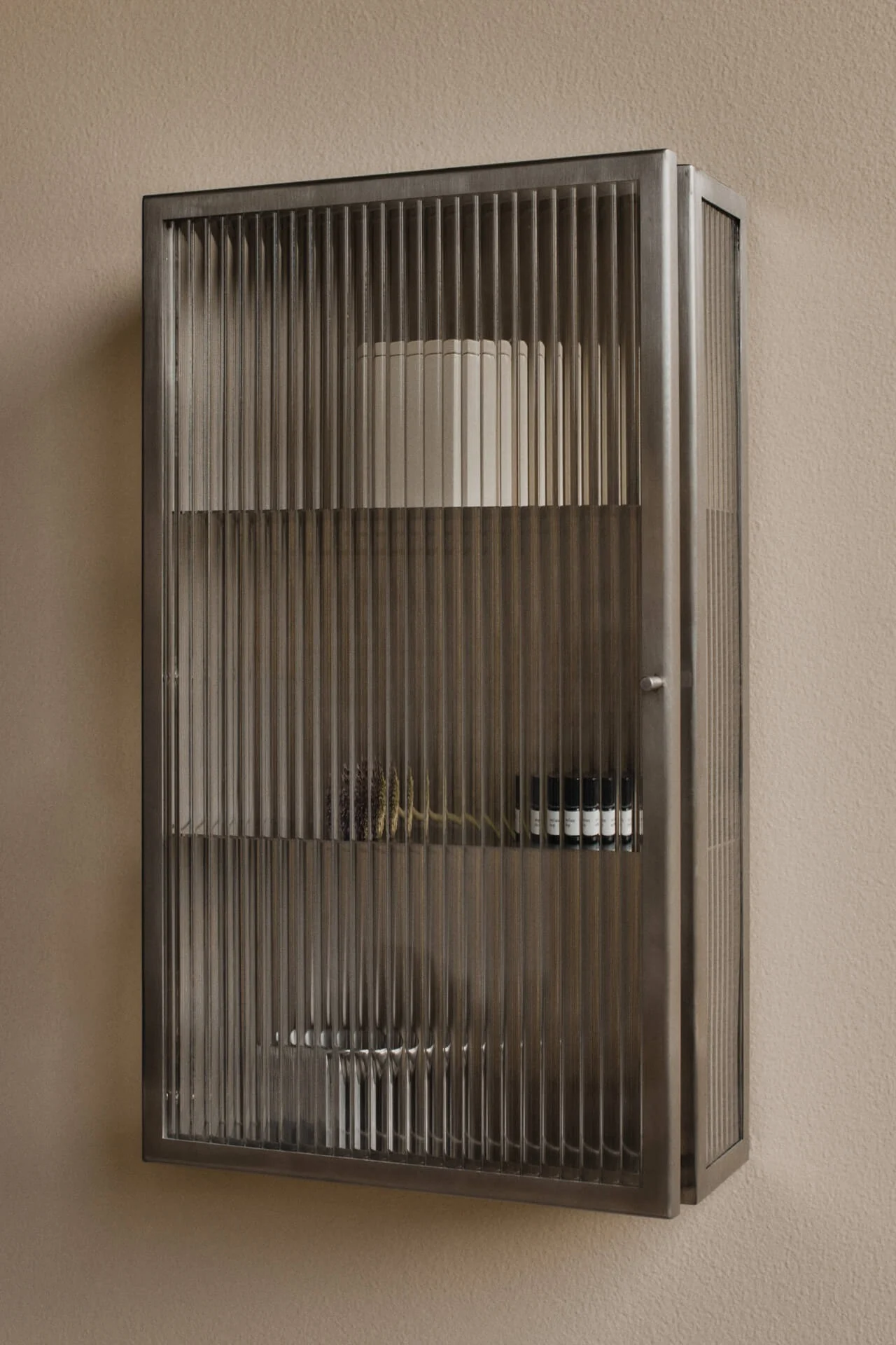 Væghængt, mørkt metalskab med riflede glasdøre på en beige væg. Indeholder hylder med en hvid cylinder, tørrede planter og små mørke flasker.