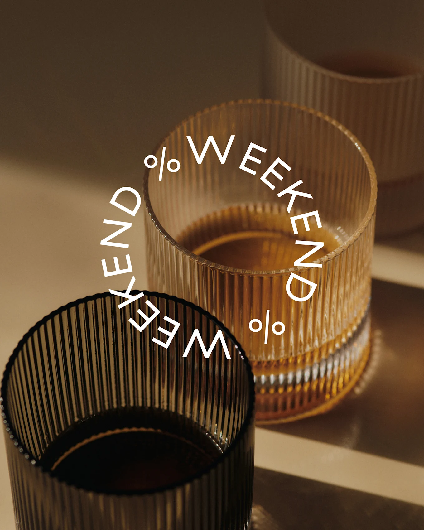 Tre riflede glas, to fyldt med mørk væske. Et mørkt glas i forgrunden, et ravfarvet glas bagved. Tekst: Weekend %.
