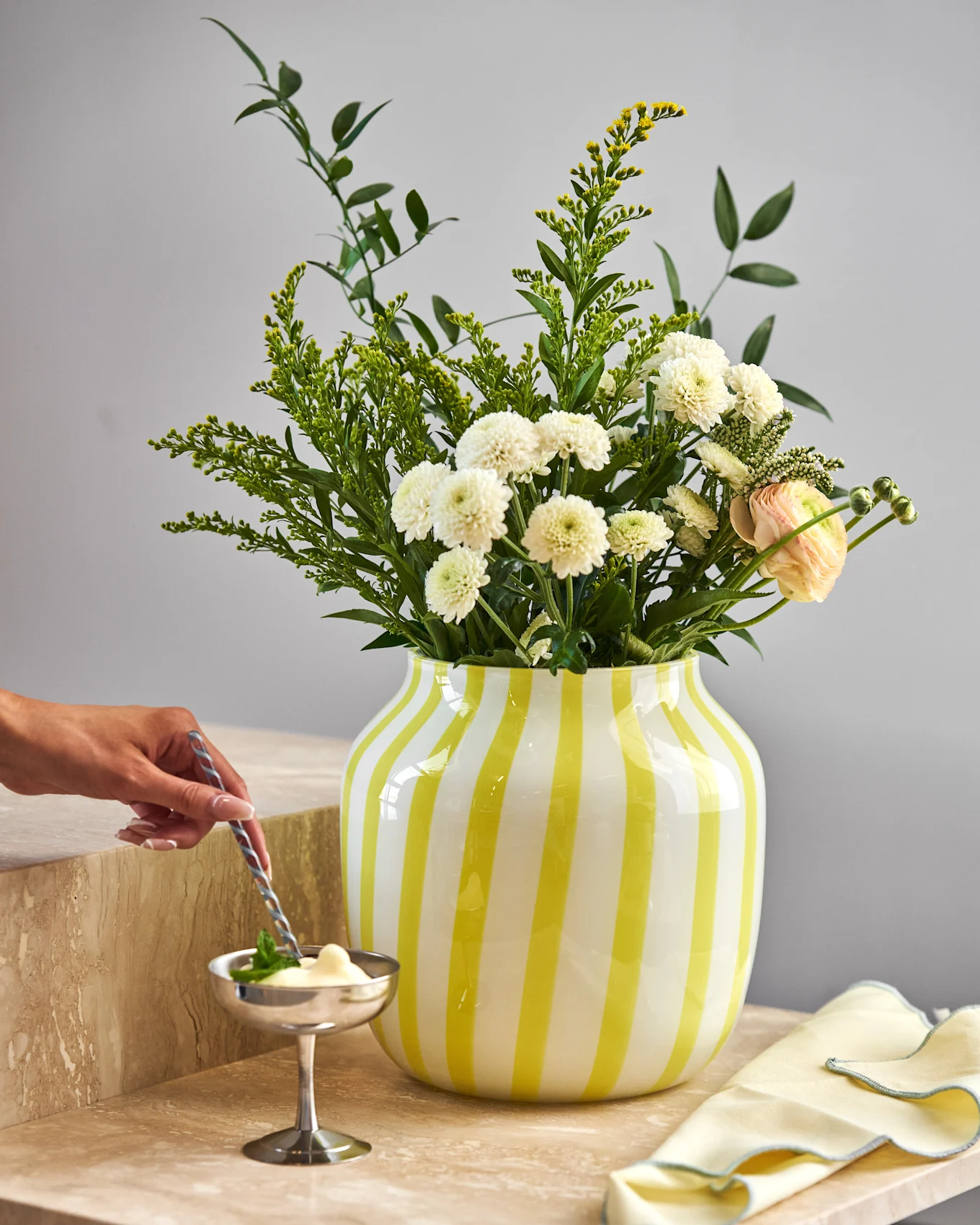 Hånd rører dessert i sølvskål på marmor. Gulstribet vase holder hvide krysantemum, gule blomster og lyserød ranunkel.