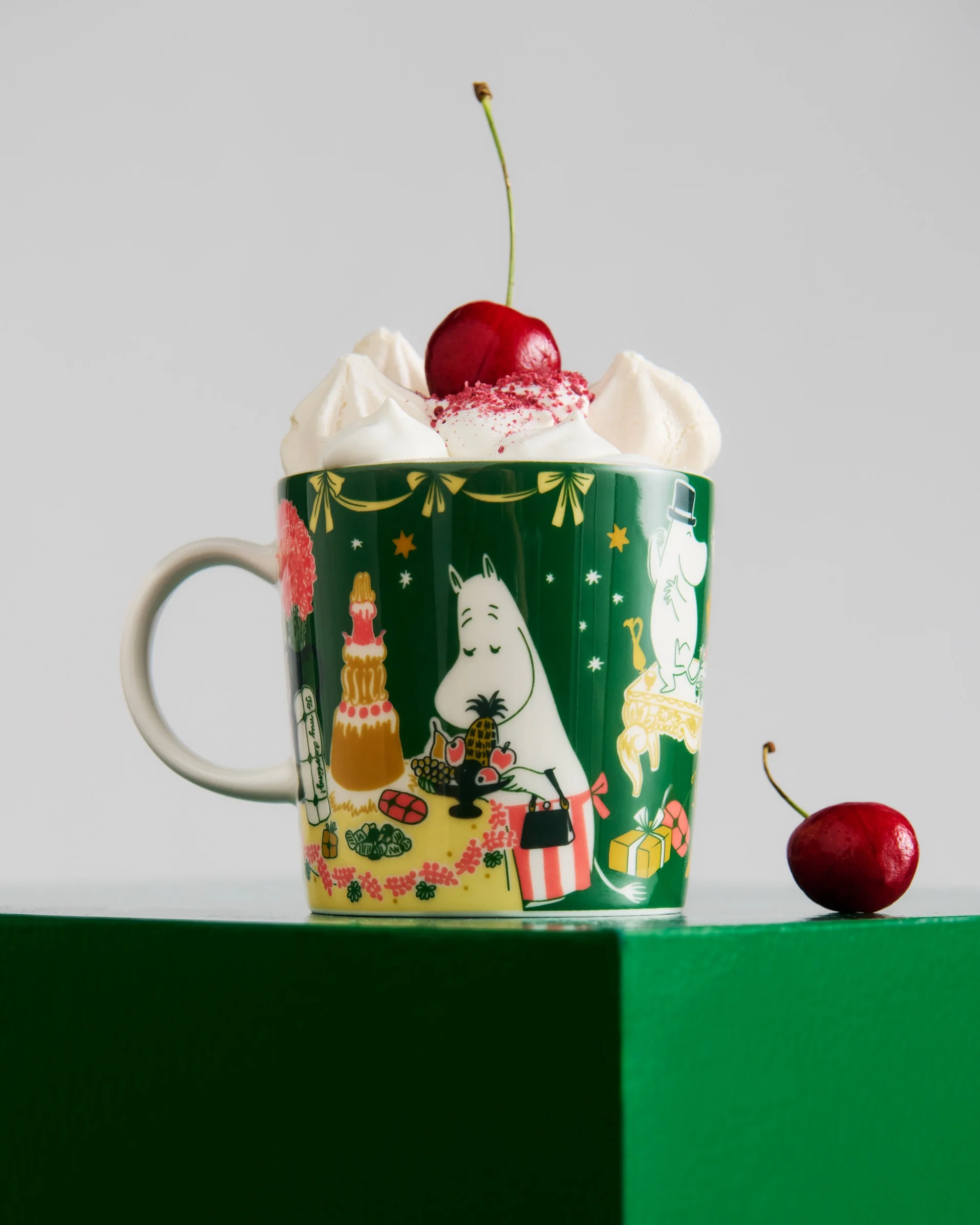 Mørkegrønt krus med Moomin-figurer, toppet med flødeskum, rødt krymmel og et kirsebær. Et andet kirsebær ligger på den grønne overflade.