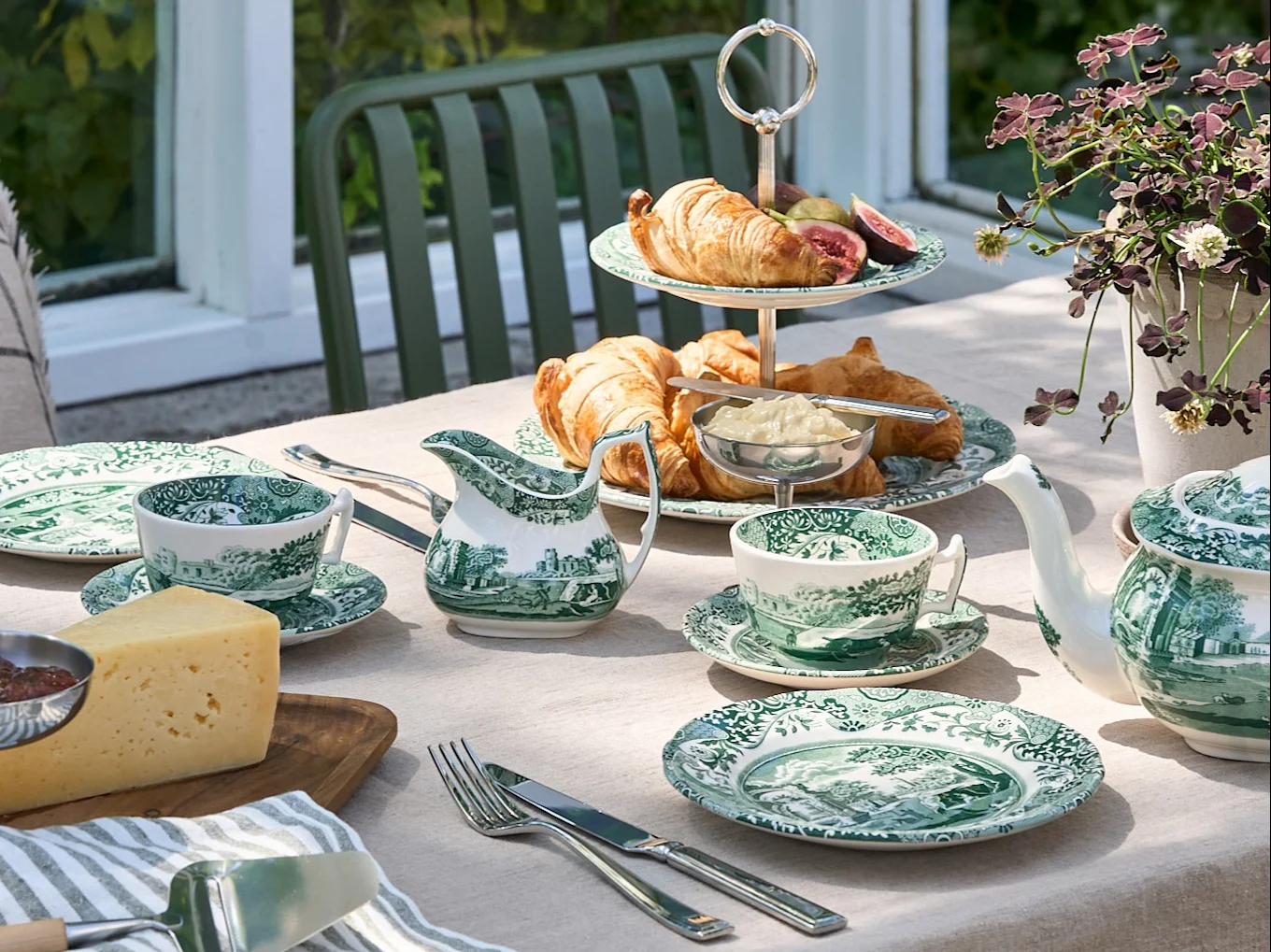 Et udendørsbord dækket med grøn-hvidt porcelæn, croissanter, ost og en tekande i en have.