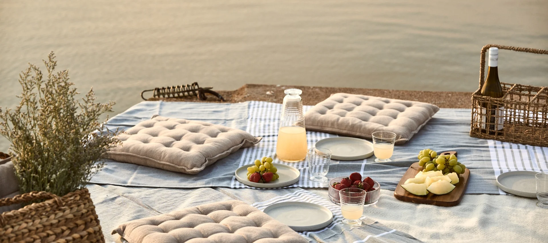 Picnic ved vandet med hynder, frugt og drikkevarer på et tæppe.