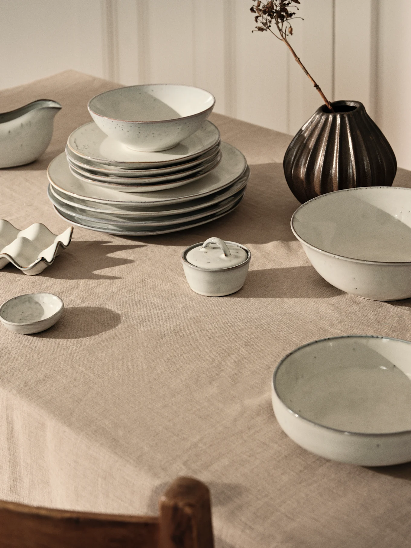 Lyst gråt keramisk stel med stablede tallerkener og skåle på beige linneddug, med mørk vase og tørret gren.