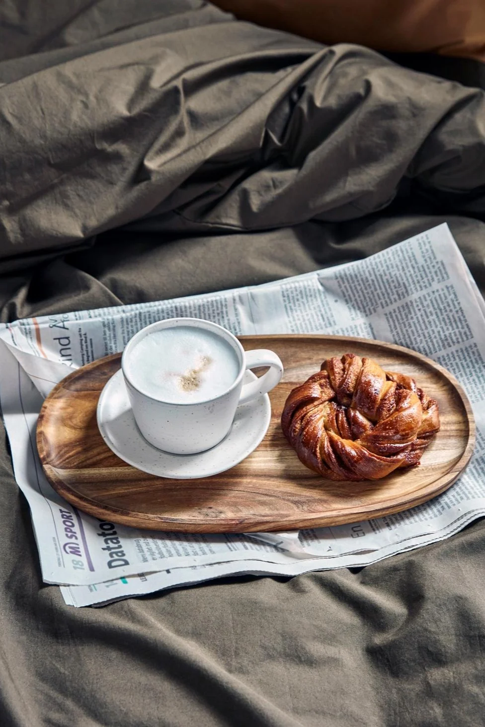 En cappuccino i House Doctors hvide og grå Pion-krus på en lille træbakke, ovenpå en avis, på en seng, med en svensk kanelbolle ved siden af.