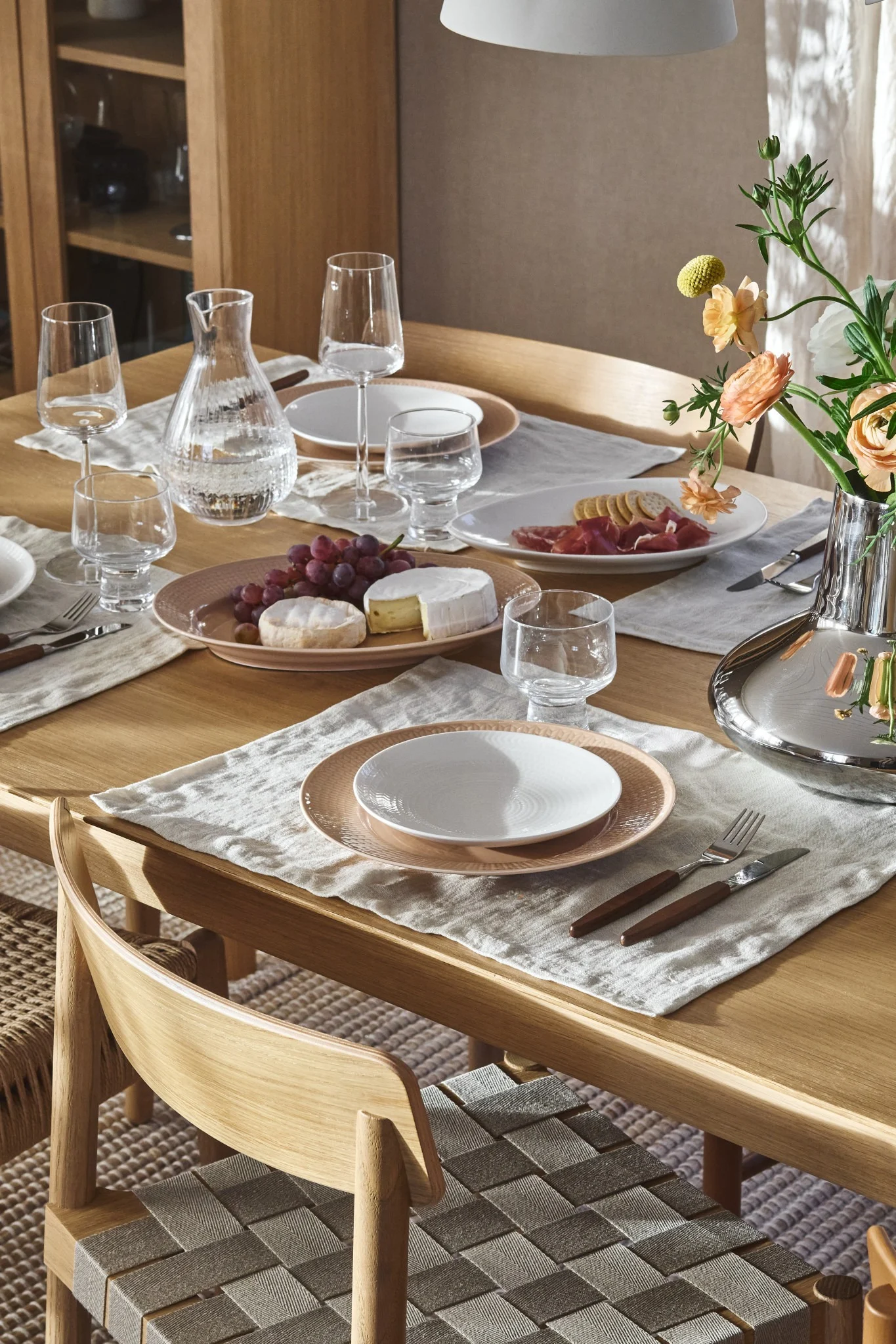 Et træspisebord dækket med tallerkener, glas, en vandkaraffel, druer, oste og pålæg, samt blomster. Flettede stole.