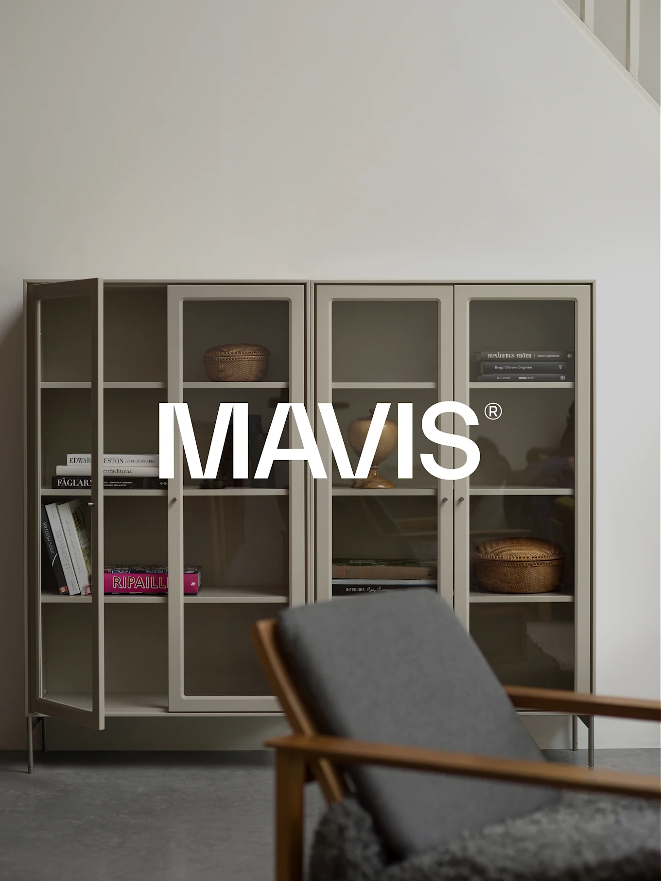 To lysegrå skabe med glasdøre, der viser bøger og indretning. En træstol er sløret i forgrunden. 'MAVIS®'-logoet er centreret.