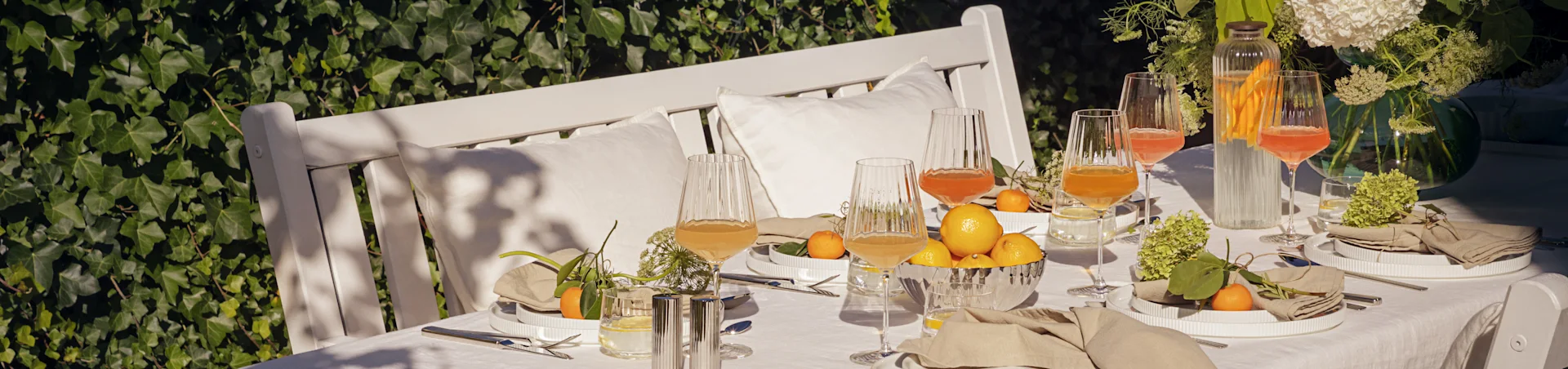 Udendørs bord dækket til sommermåltid med hvide ribbede tallerkener, linnedservietter, riflede glas, citroner og mandariner.