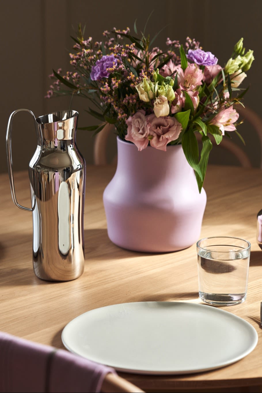Her ses den lilla Dorotea-vase med en buket lyserøde og lilla blomster stående på et bord ved siden af den sølvfarvede Dorotea-karaffel.