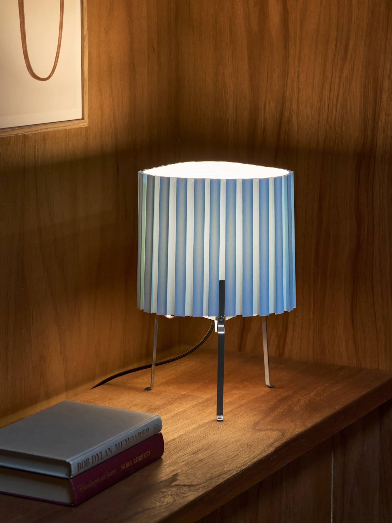 En blå- og hvidstribet plisseret bordlampe med tre metalliske ben står på en træhylde ved siden af to bøger. Indrammet kunst i baggrunden.