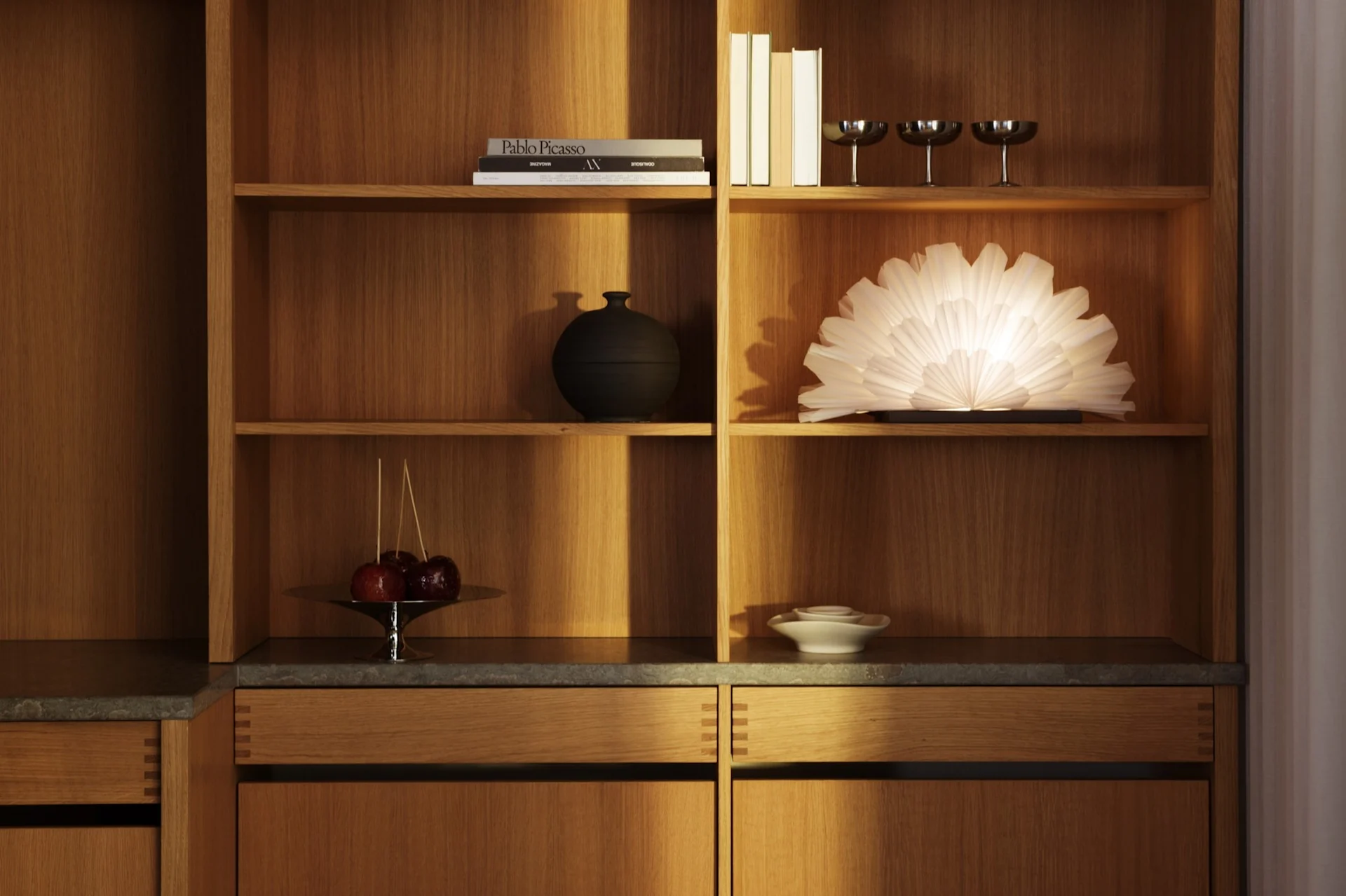 Et moderne trækabinet med hylder, der viser bøger, en sort vase, tre sølvglas, en glødende vifteformet lampe, kandiserede æbler og hvide keramiske skåle.