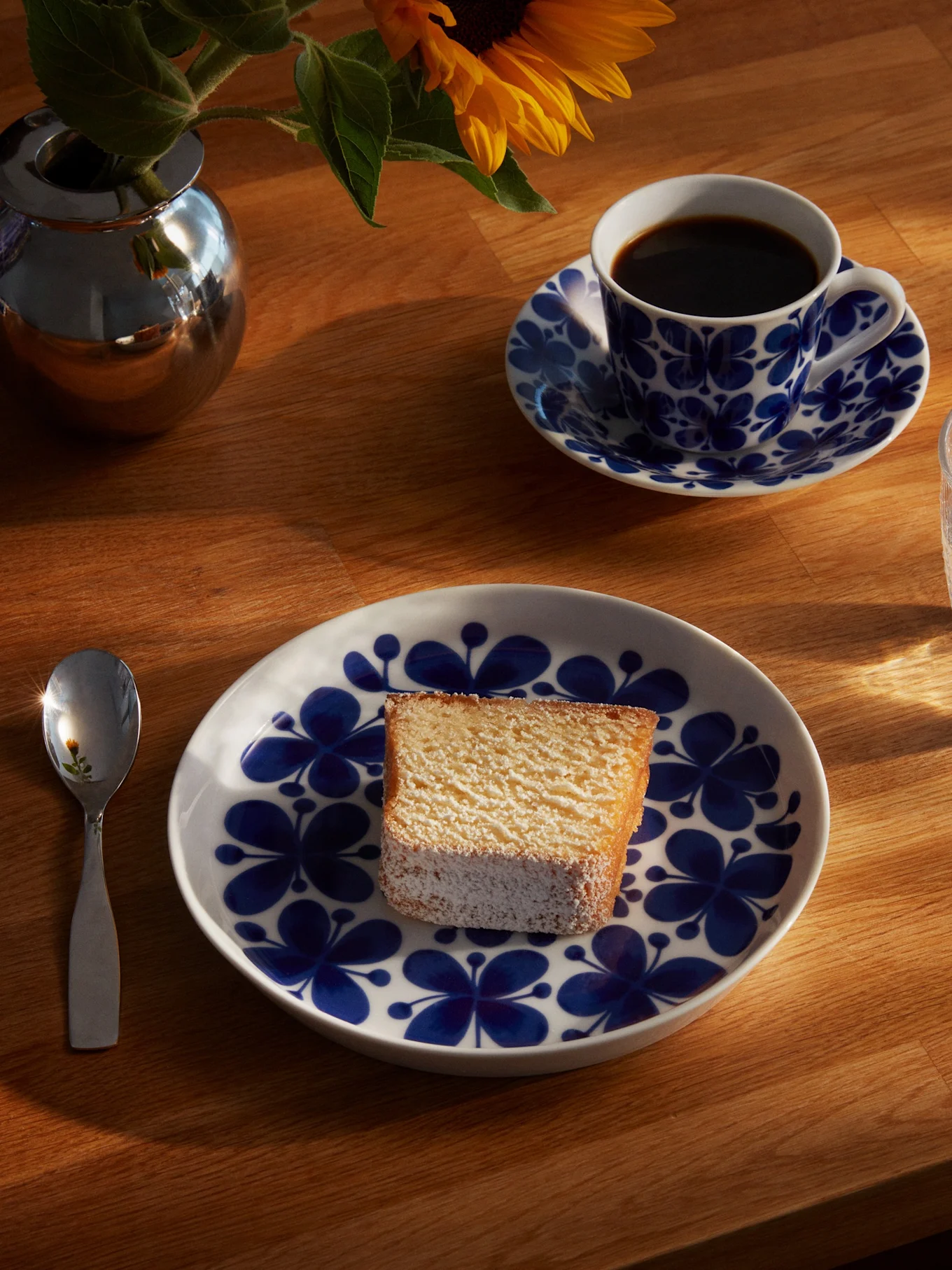 Et stykke kage på en blå og hvid blomstret tallerken, med en kaffekop, et glas vand, en ske og en solsikke i en sølvvase på et solbeskinnet træbord.