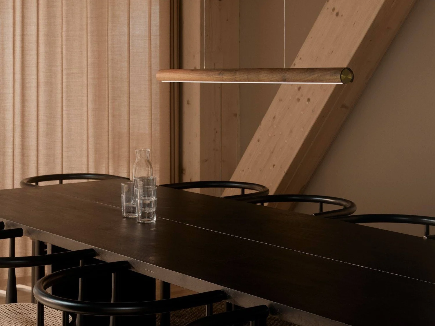 Et moderne spiseværelse med et langt mørkt træbord, sorte stole og en slank pendellampe i træ. En glaskaraffel og tre glas står på bordet.