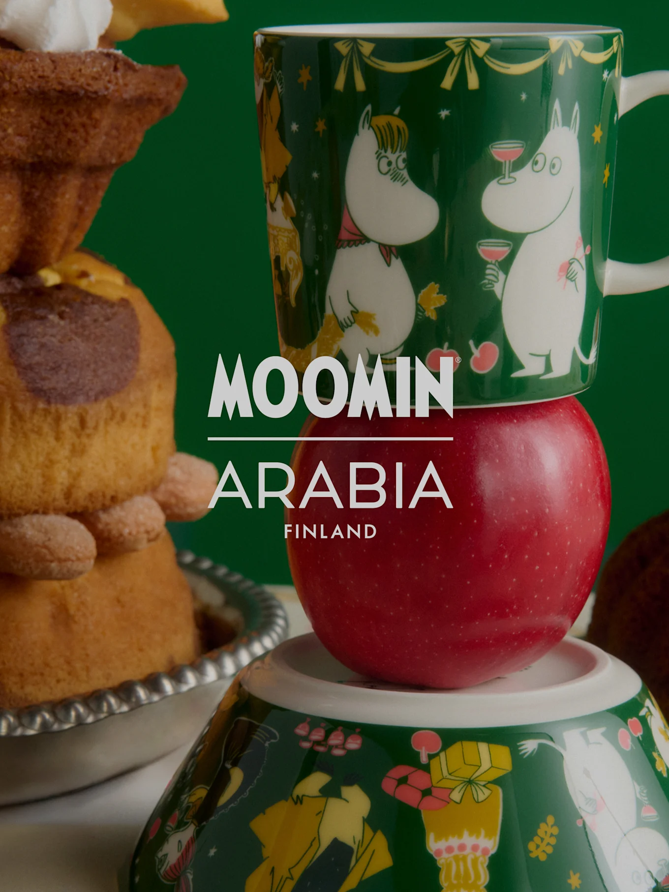 Moomin Arabia julekollektion: Grønt Mummi-krus på et rødt æble, oven på en Mummi-skål, ved siden af festlige kager.