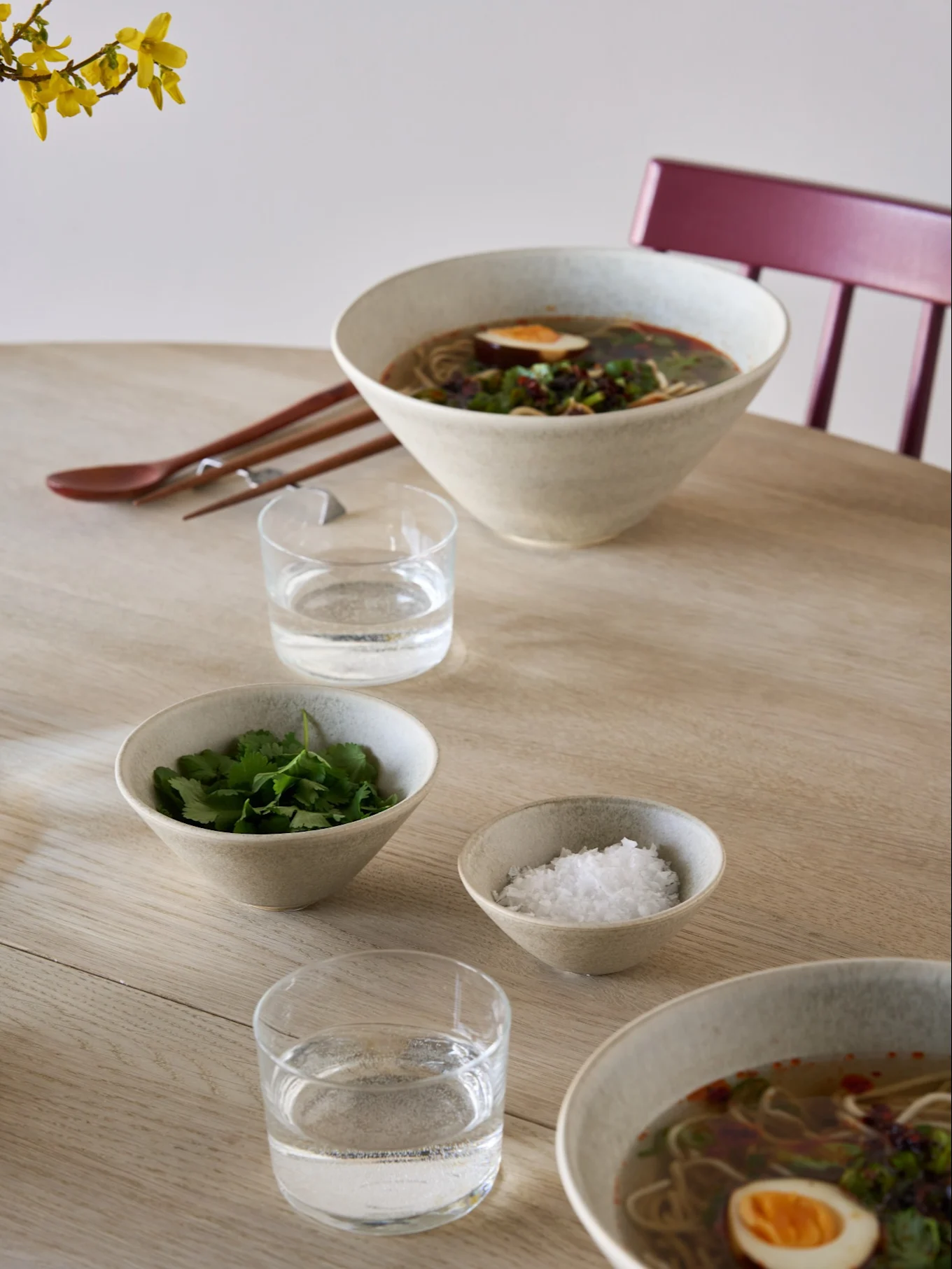 To skåle ramen med æg og urter, sidetallerkener med koriander og salt, vand, spisepinde og en ske på et lyst træbord.