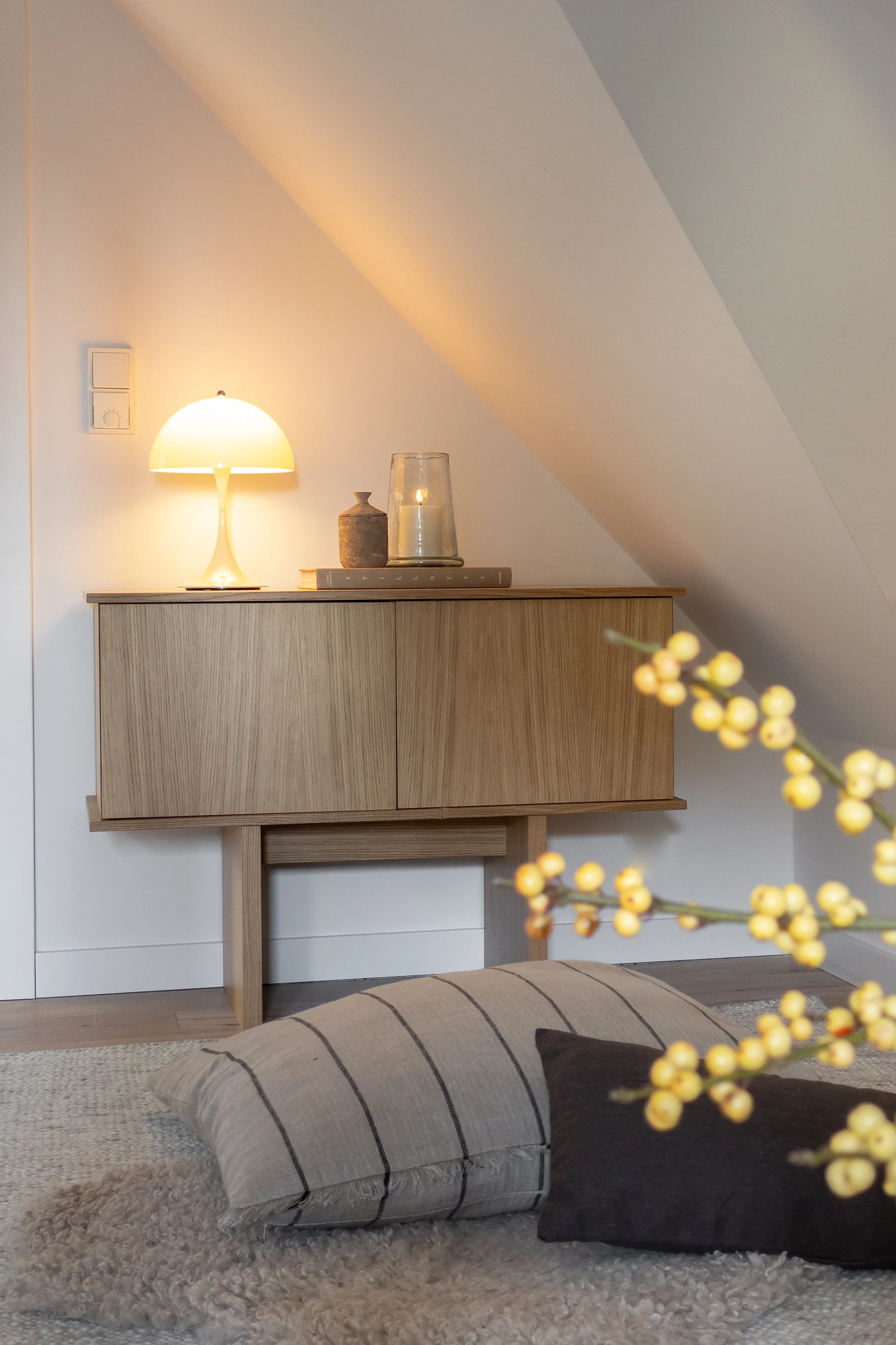 Et træskab med en glødende champignonlampe og et tændt lys, mod en hvid, skrå væg i et hyggeligt rum.