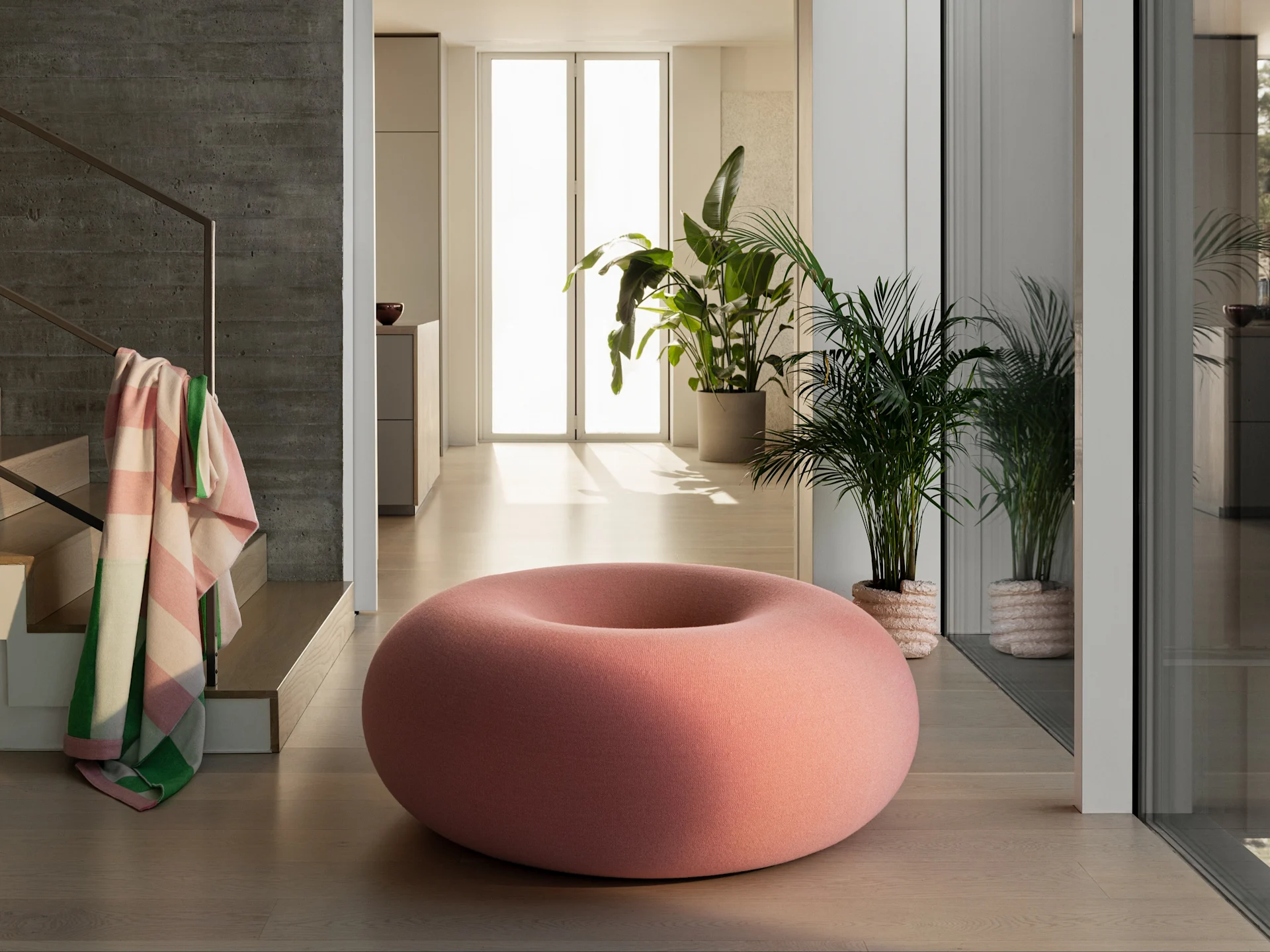 En stor, donutformet pink puf på et lyst trægulv i et moderne rum med grønne planter og en betonvæg.