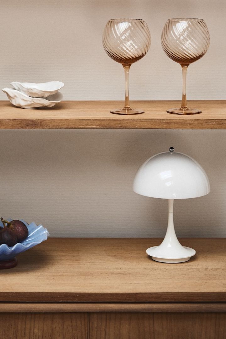 Forny dit hjem med moderne belysning som Panthella mini fra Louis Poulsen, der her ses i et vitrineskab af træ med pink vinglas og en blå skål. 