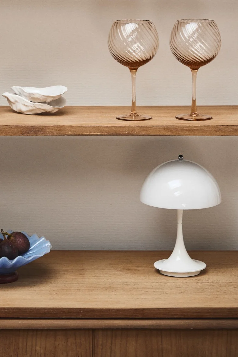 Forny dit hjem med moderne belysning som Panthella mini fra Louis Poulsen, der her ses i et vitrineskab af træ med pink vinglas og en blå skål.