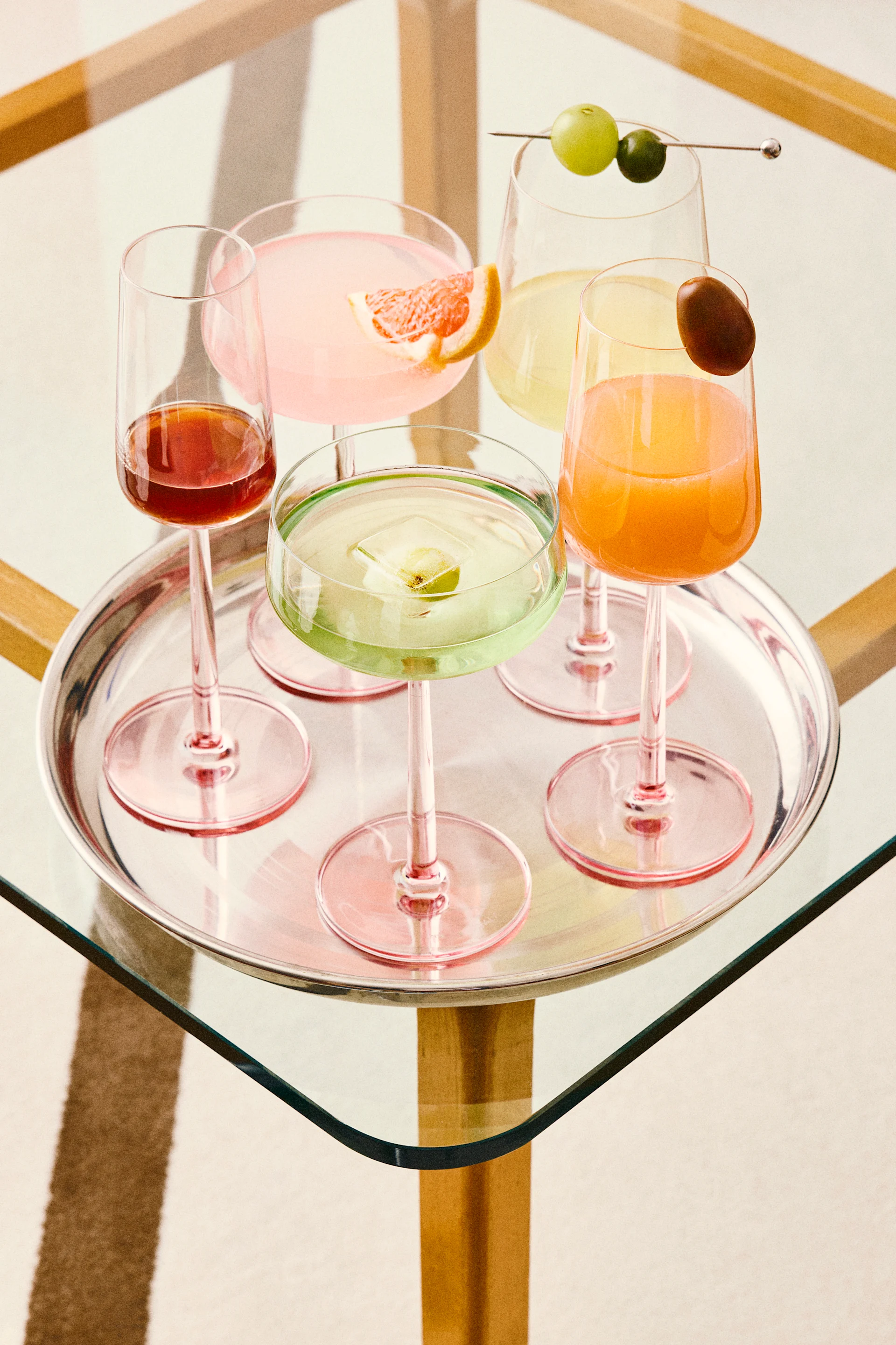 Fem farverige cocktails på en sølvbakke, inklusiv pink, grønne, orange og røde drinks med diverse garniturer, på et glasbord.