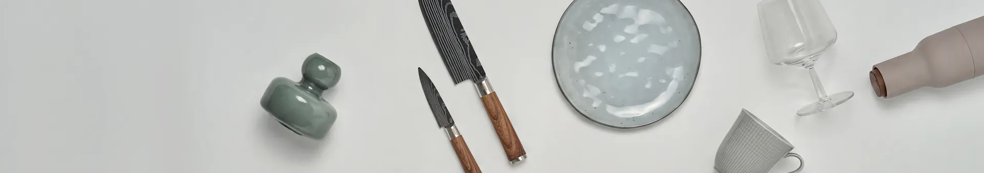 Diverse køkken- og spisestel, herunder en grøn vase, to knive, en plettet tallerken, et vinglas, en beige flaske og et mønstret krus.