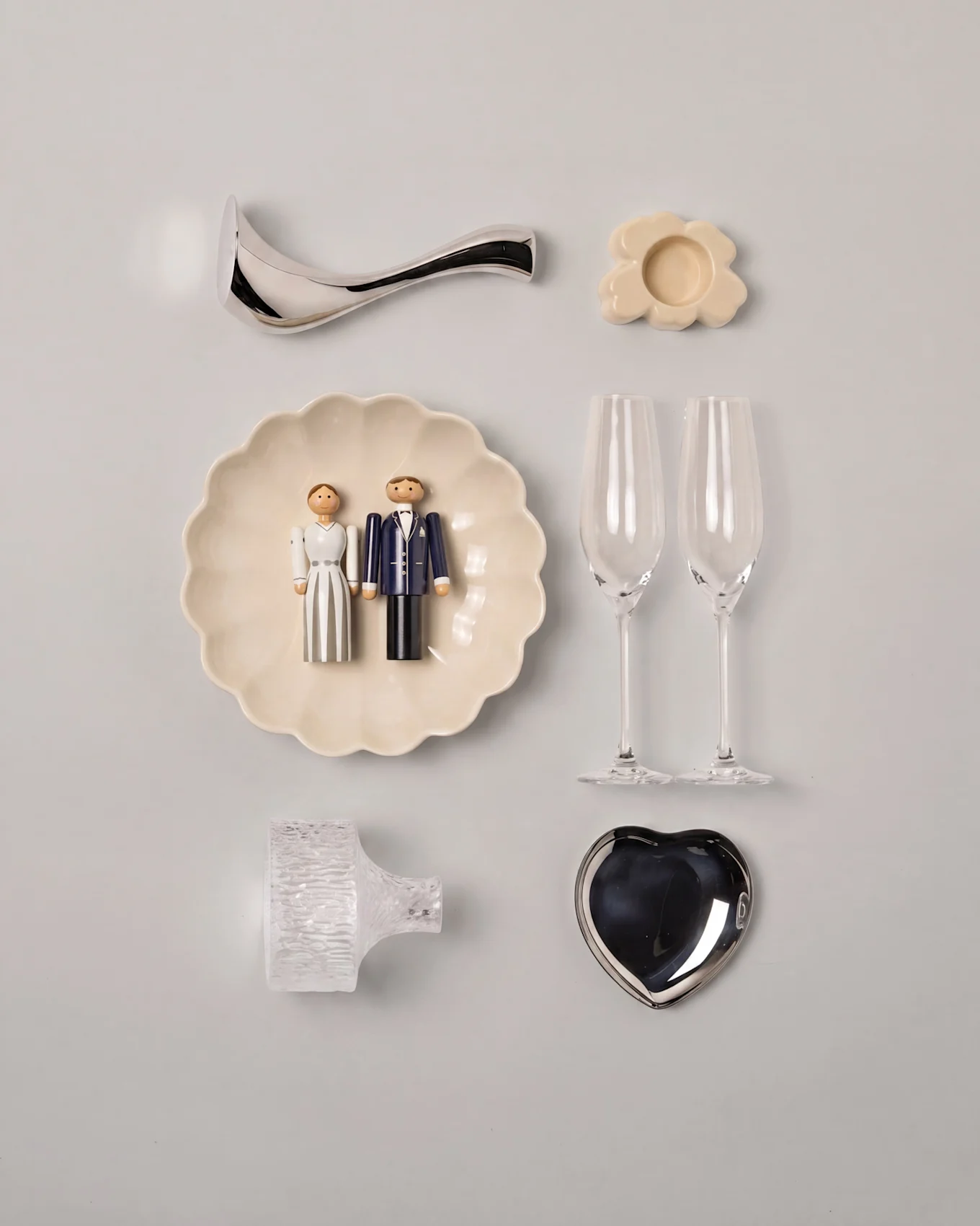Bryllups-flat lay med træ brude- og gomfigurer på en muslingeskål, to champagneglas, et sølv hjertefad og assorteret pynt.