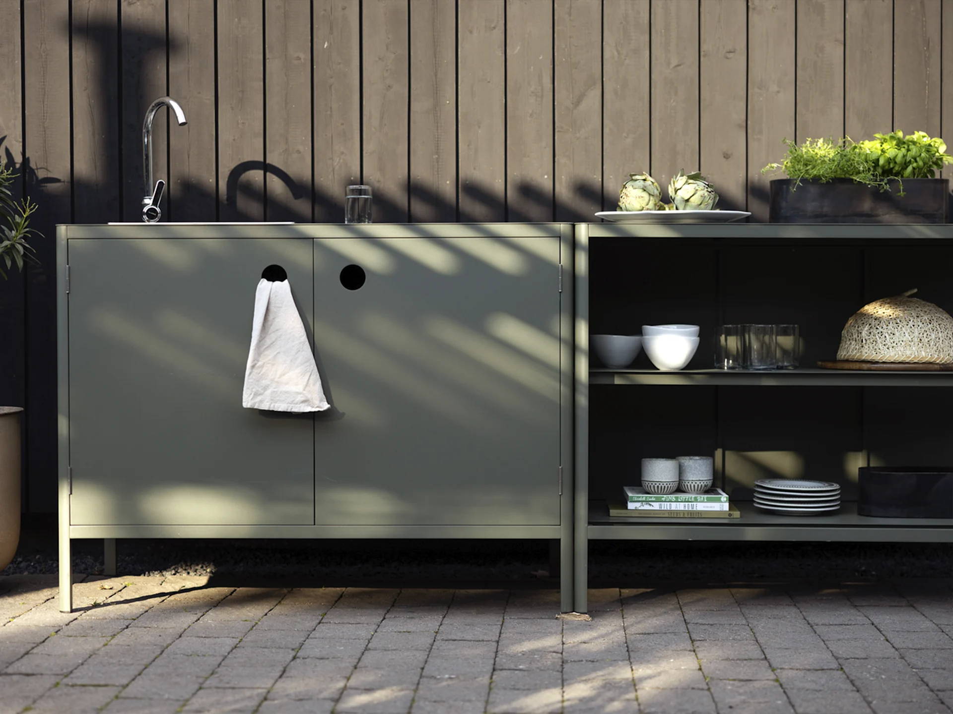 Udendørskøkken med mørkegrønne skabe, en vask, vandhane og hylder med artiskokker, urter og service.