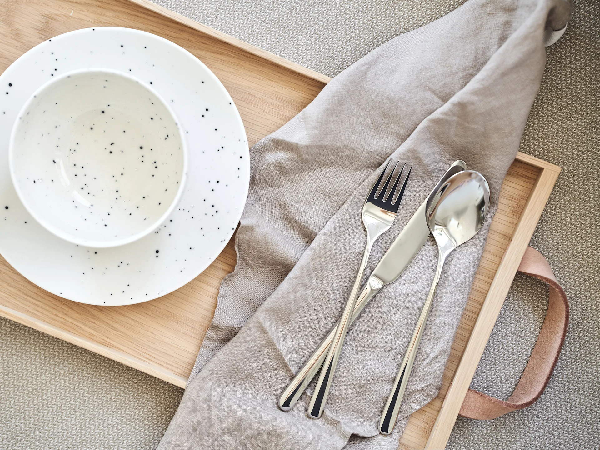 En træbakke med en hvid, plettet skål og tallerken, en grå linnedserviet og sølvbestik på en vævet måtte.