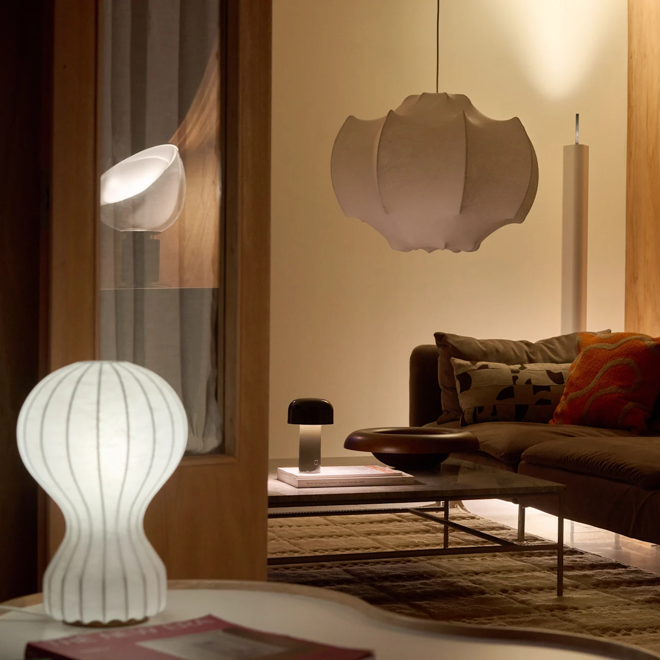 En stue med en brun sofa og sofabord, oplyst af en hvid riflet bordlampe, en mørk svampelampe og en stor plisseret pendellampe, der skaber en varm glød.