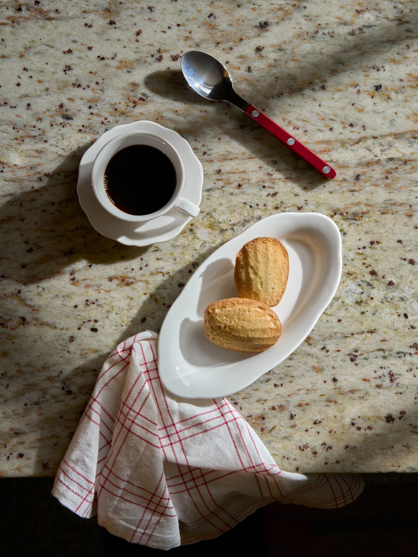 Ovenfra af sort kaffe i hvid kop, to madeleinekager på hvid tallerken, ske og stribet serviet på marmorbord.