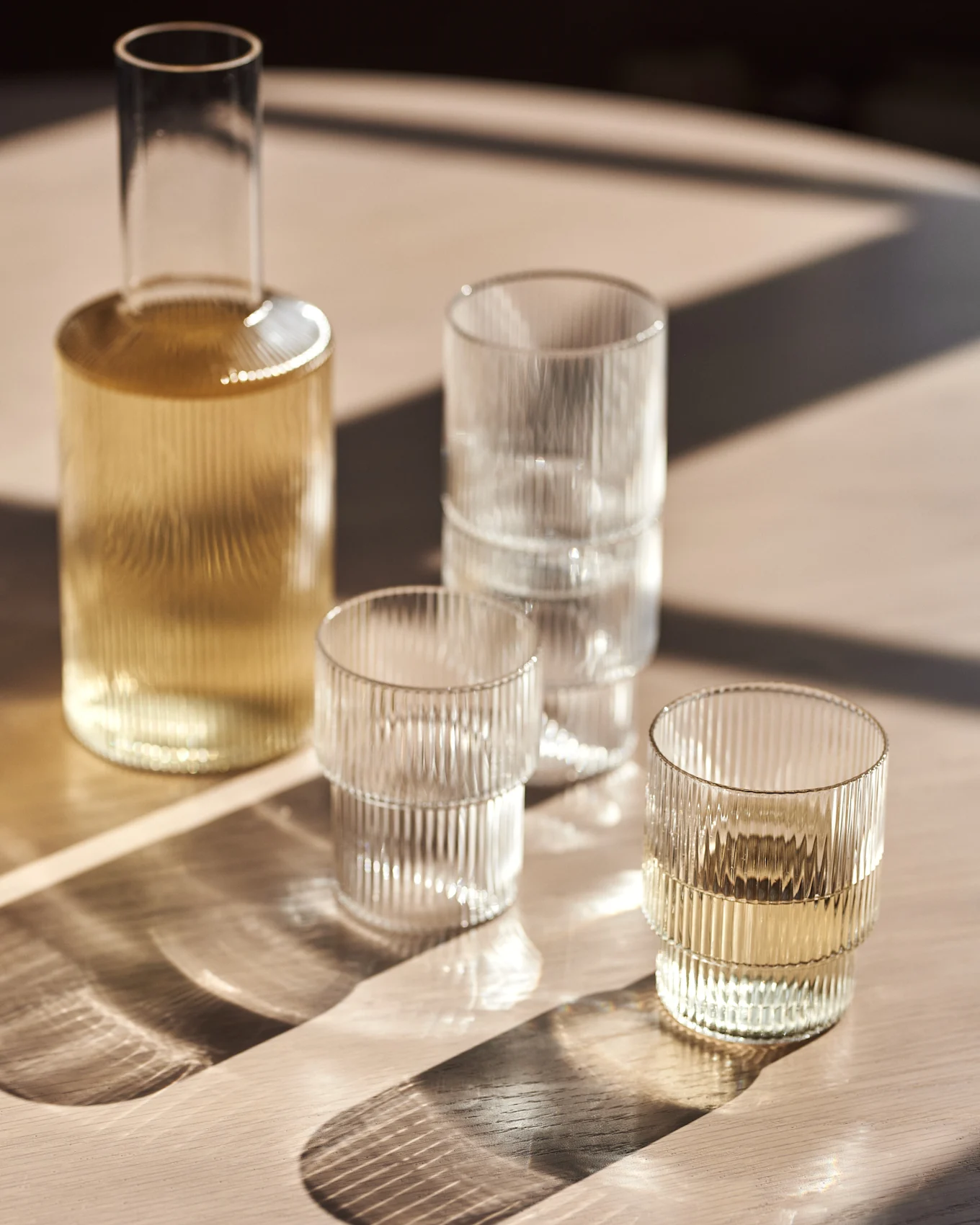 Glaskaraffel og riflede glas på et træbord med sollys.