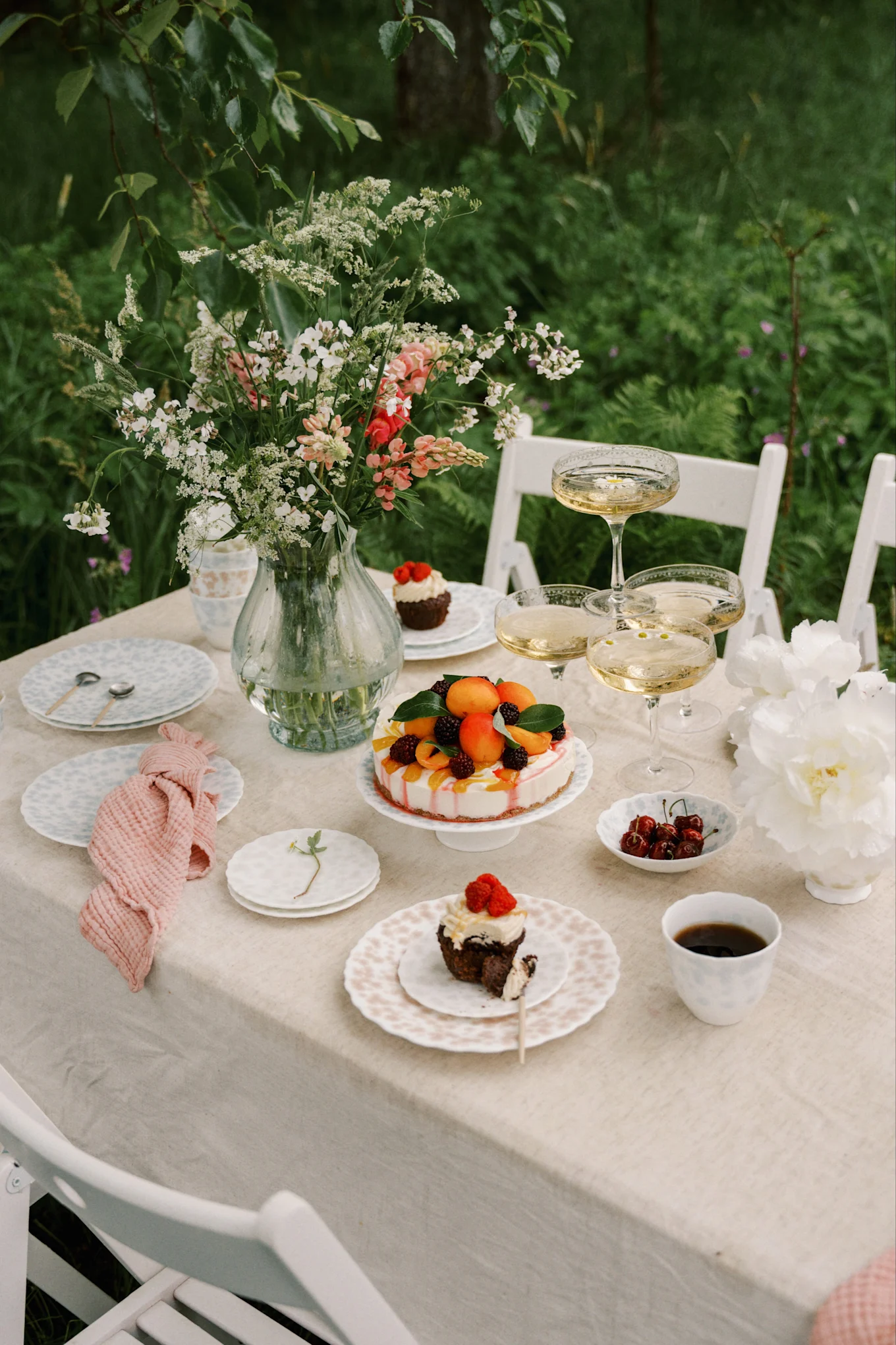 Udendørs bord dækket med blomstervase, champagne, frugtkage og cupcakes med bær.