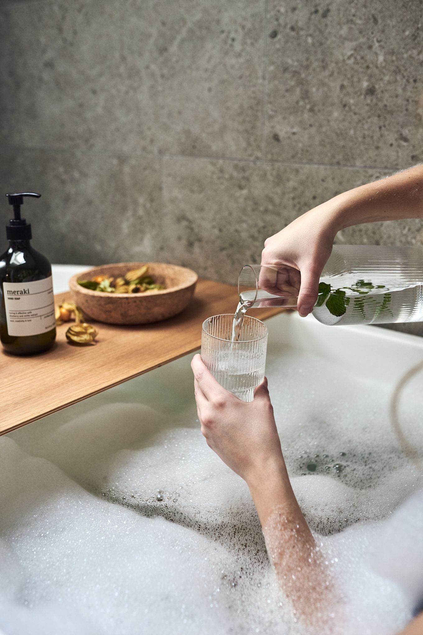 Hænder der hælder vand i et Ripple glas fra ferm LIVING som en del af hjemmespaen i et varmt boblebad.