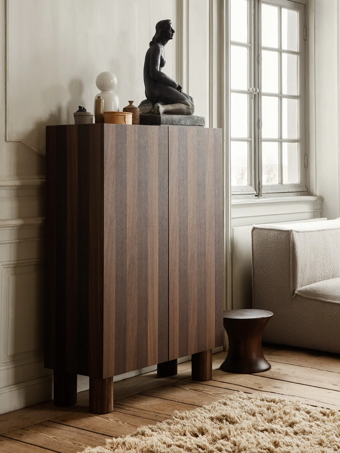 Mørkt træskab med lodrette striber, toppet af en siddende skulptur, hvid lampe og indretning, ved siden af en tekstureret sofa og mørk skammel.