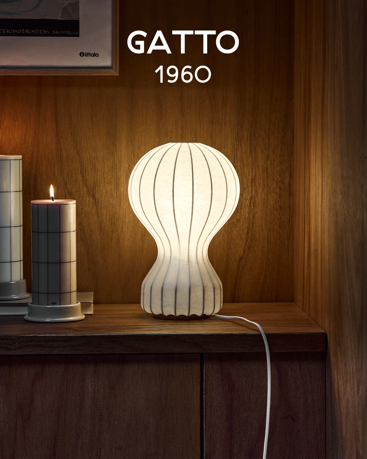 En lysende hvid, ribbet lampe med timeglasform står på en træhylde ved siden af et tændt stearinlys. Tekst: GATTO 1960.