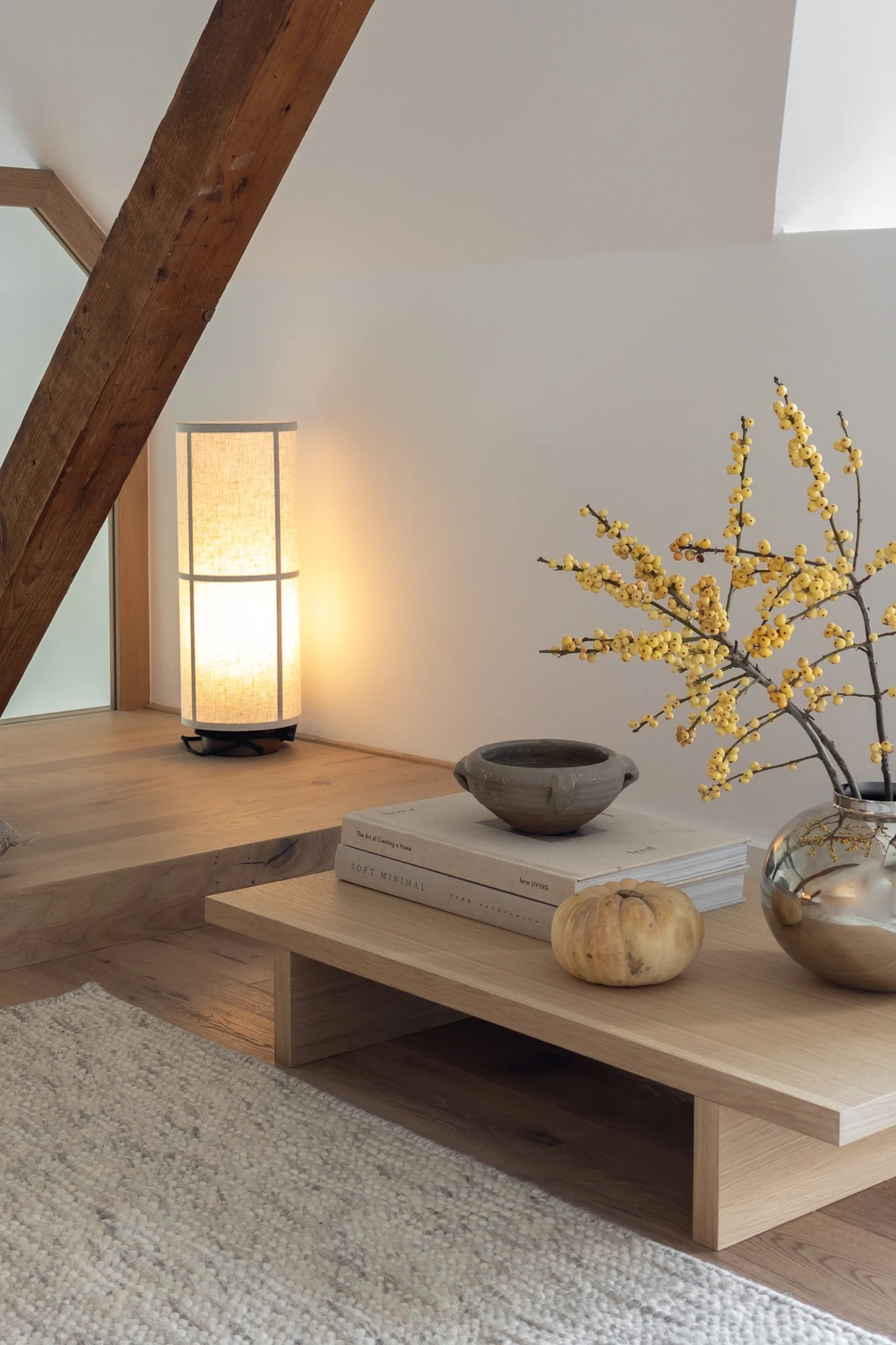 Hyggelig loftstue med høj lampe, træbjælke, lavt sofabord med gule bær, bøger og et lille græskar.