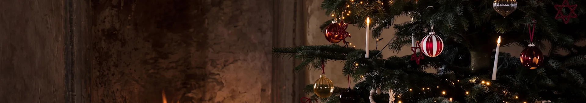 Et festligt juletræ med pynt og et tændt lys ved siden af en varm pejs med brændende brænde.