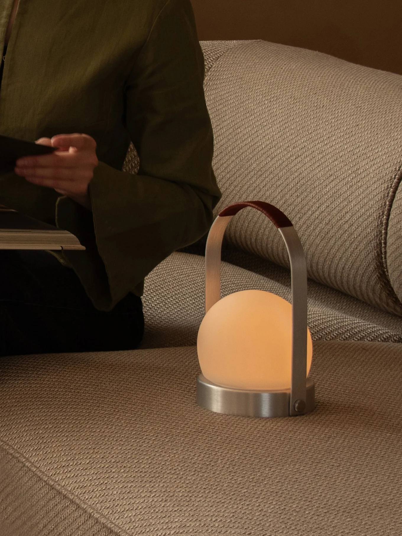 Person læser på en sofa med en glødende bærbar lampe med matteret kugle, metalbase og læderhåndtag.