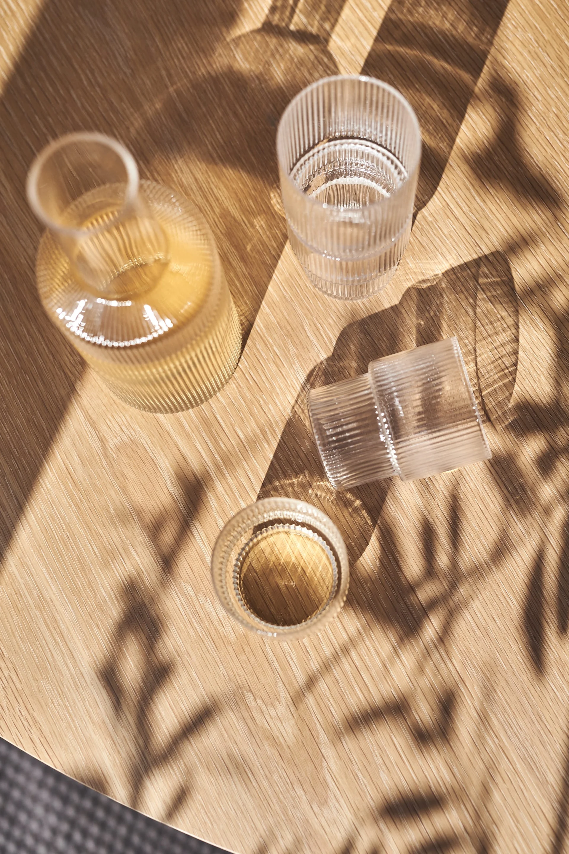 En riflet glasflaske og to riflede glas, det ene med væske, på et solbeskinnet træbord med bladskygger.