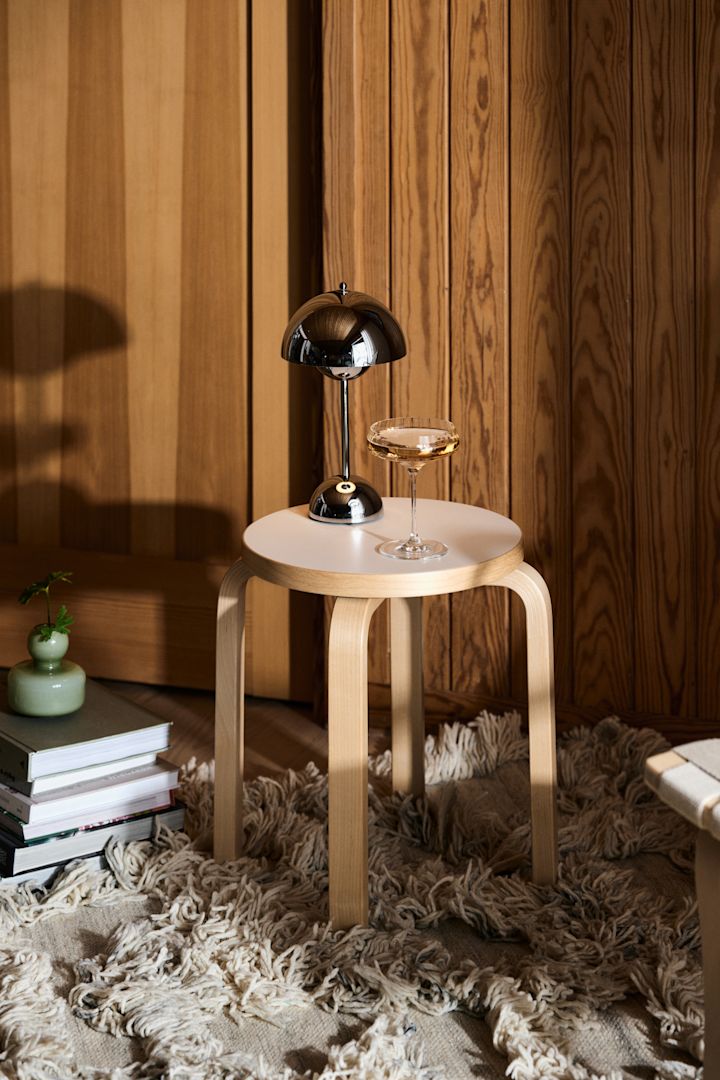 En af efterårets store trends 2024 er krom. Her ser du den klassiske bærbare lampe FlowerPot VP9 i krom fra &Tradition, stående på en skammel ved siden af et glas champagne.