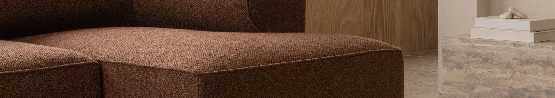 Nærbillede af en brun tekstureret sofa, med en trævæg og dekorative genstande på en betonoverflade i baggrunden.