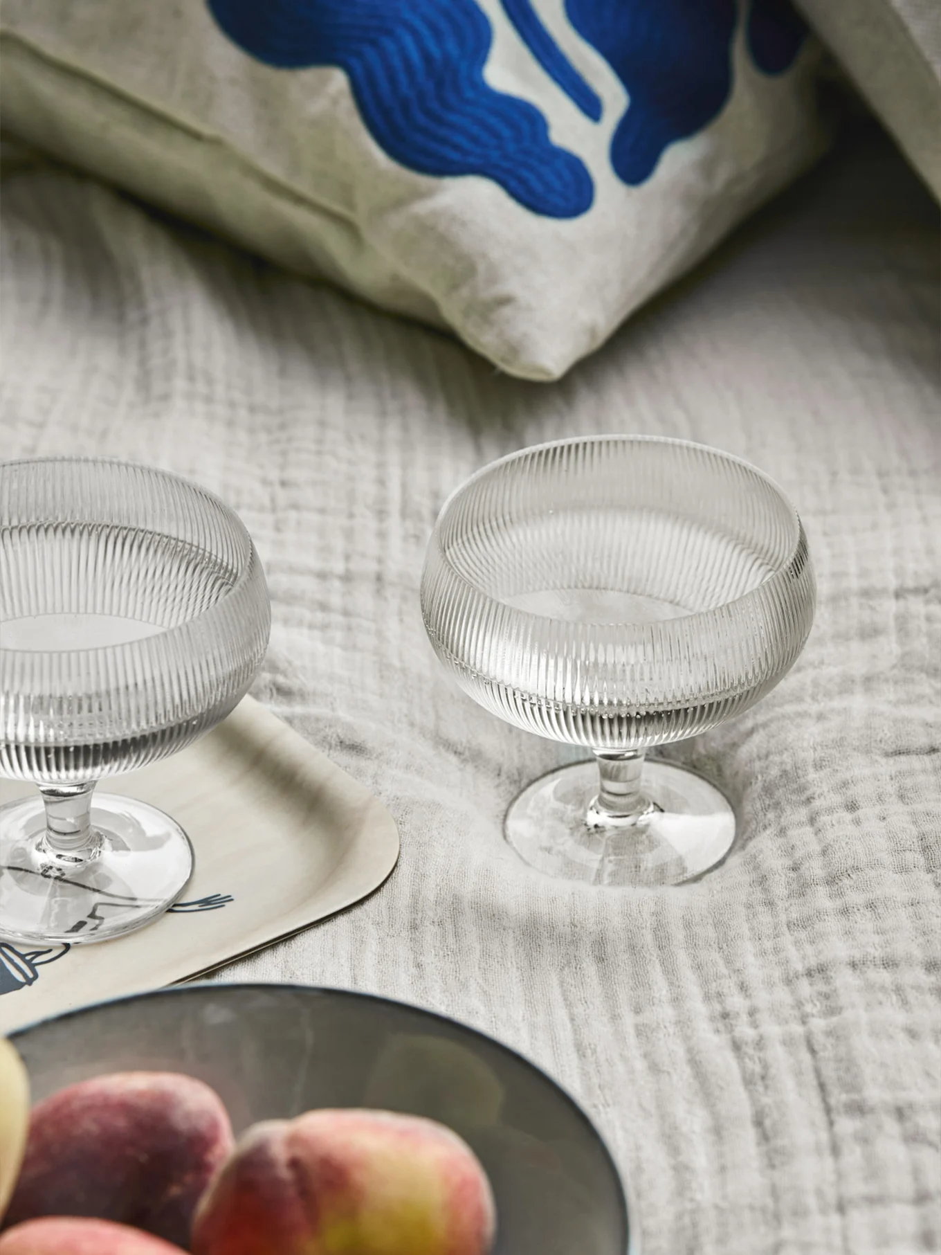 To riflede glasdessertskåle, en pude med blå abstrakt kunst, en træbakke og ferskner på et lyst stof.