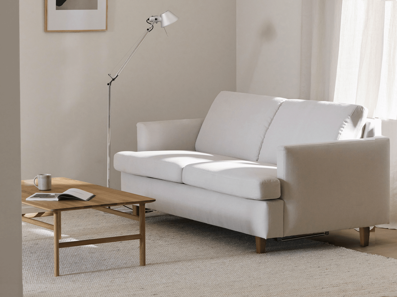 En minimalistisk stue med en lysegrå to-personers sofa, et sofabord i træ, en moderne gulvlampe og gennemsigtige gardiner.