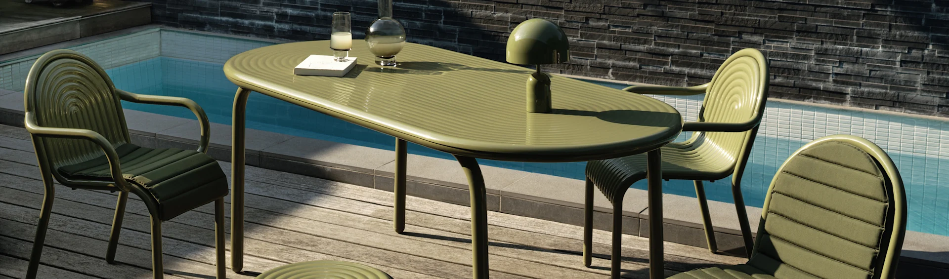 Grønt ribbet udendørs spisebord og stole på en træterrasse ved en pool. En lampe, karaffel, glas og bog er på bordet.